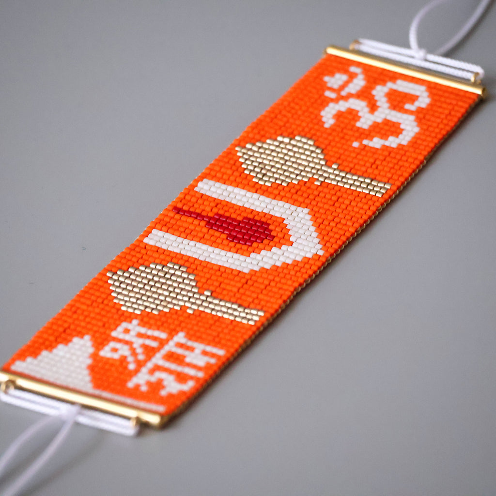 A handmade beaded bracelet featuring a bright orange base with white and red beads forming the Om symbol, a lotus flower, and Sanskrit mantras, finished with gold-toned metal end caps and white cord ties.